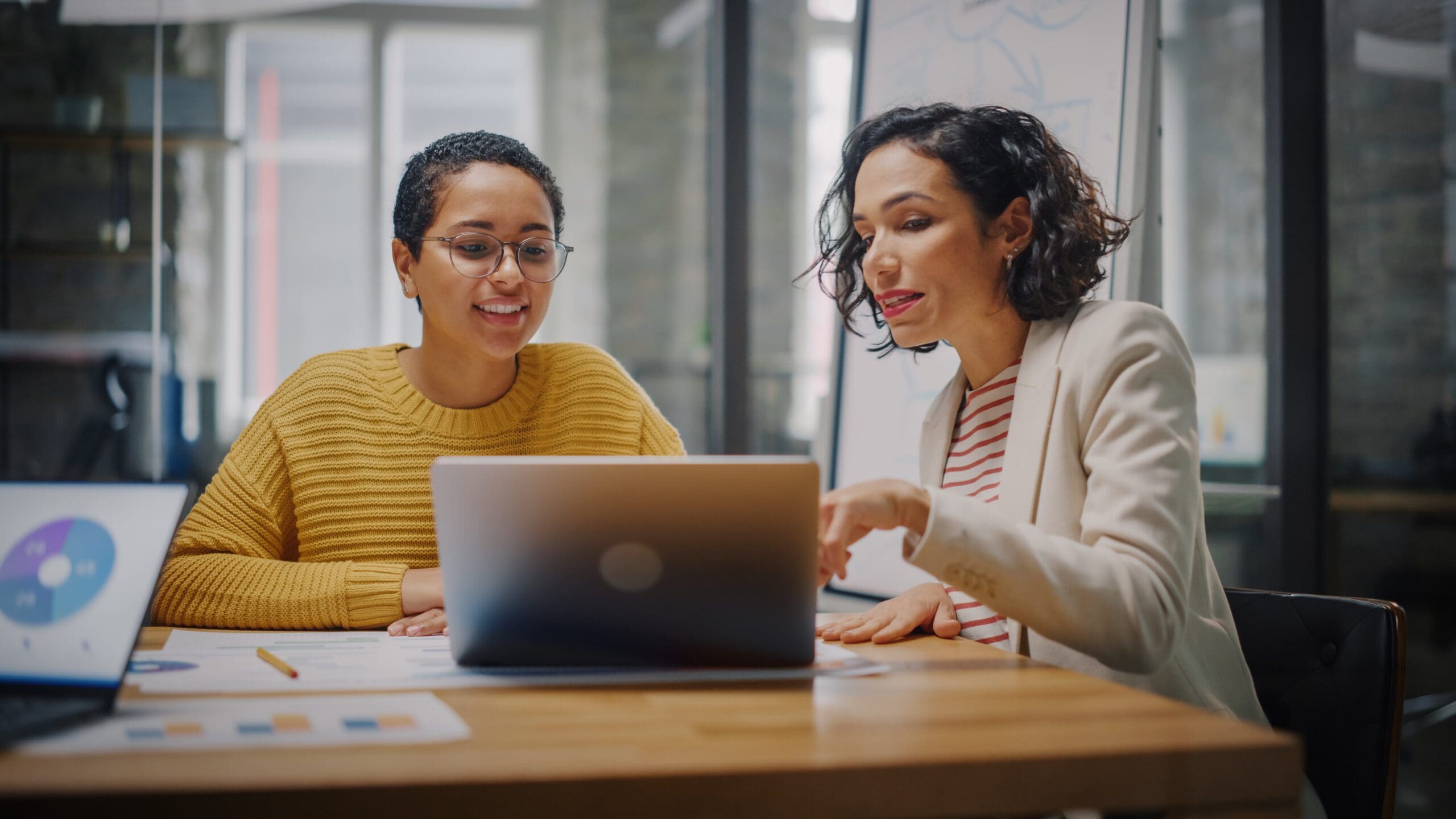 Two women have a discussion in a meeting room while looking at a laptop